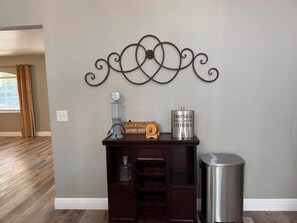 A room with a wall-mounted decorative metal piece, a wooden cabinet with a sign, a metal container labeled 'Drinks Well with Others', and a trash can. - Fireline's Ocala Retreat Near WEC, Springs Hot Tub! (Ocala City)