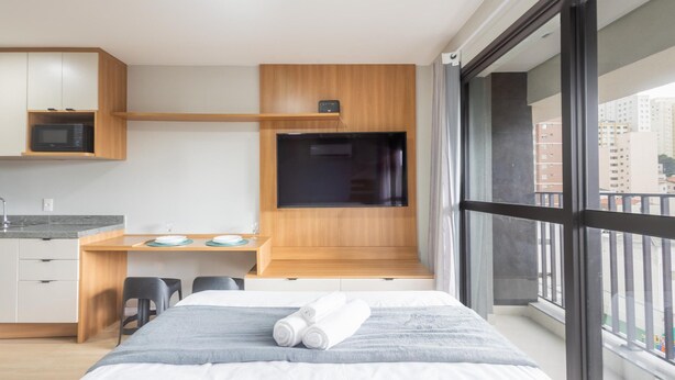 A modern hotel room with a bed, a desk, a microwave, and a large window. - Complete Studio in Perdizes Cep00428 (São Paulo)