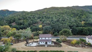 Una casa a due piani con vialetto, circondata da alberi e colline.