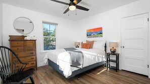 Ein Schlafzimmer mit Bett, Kommode, Stuhl, Fenster, Deckenventilator, einem Bild und einer Tür.