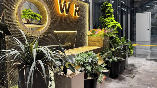 A reception area with a modern design, featuring a reception desk, a circular planter with a tree, and various potted plants. - Wind Rose hotel (Almaty)