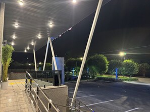 Un espacio exterior con un corredor cubierto, estacionamiento y una calle con farolas.