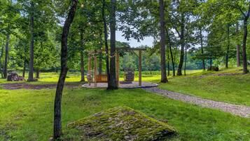 Ein friedlicher Park mit einer hölzernen Schaukel, einer Feuerstelle aus Stein und einem moosbewachsenen Baumstamm.