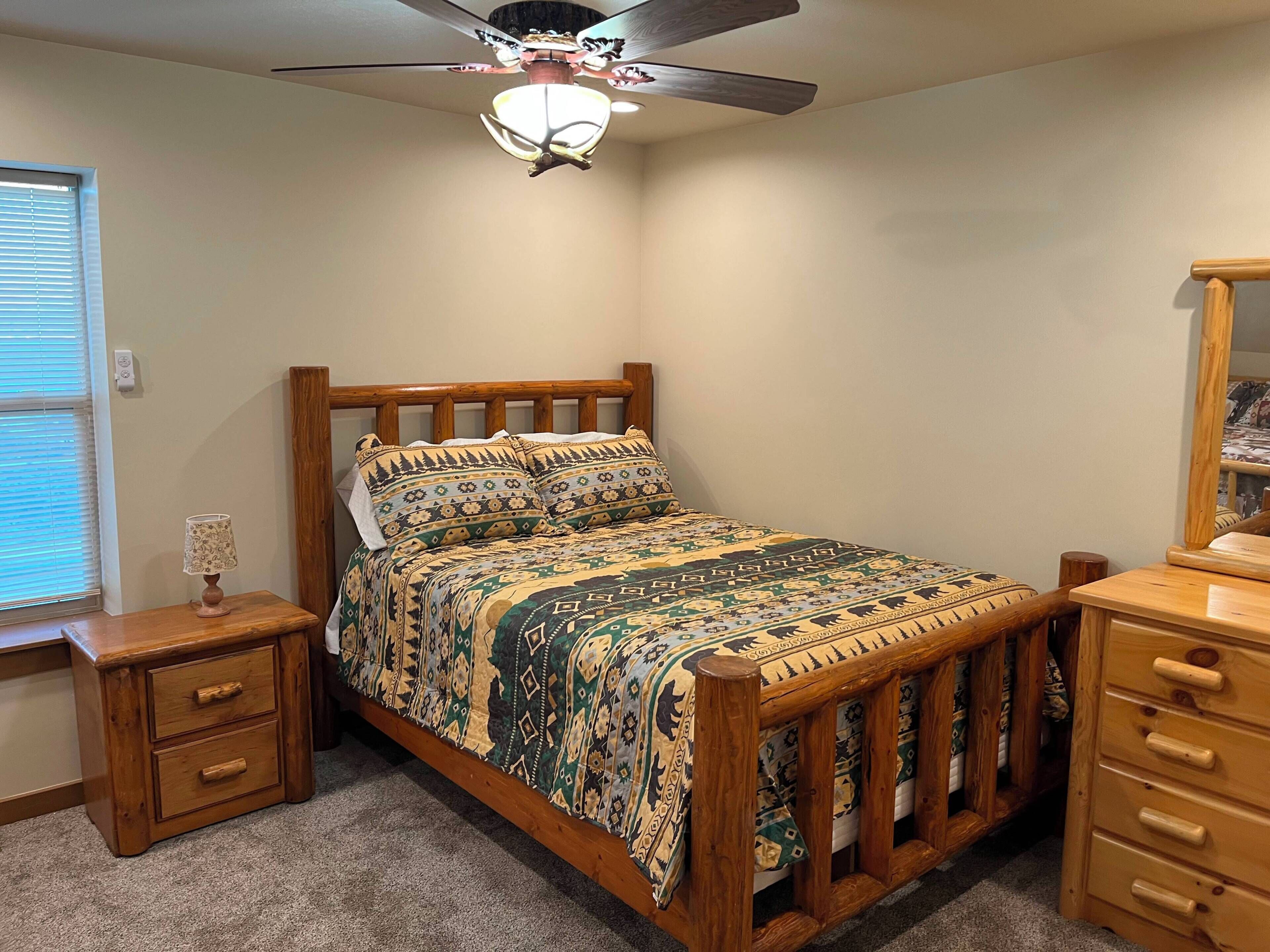 Un dormitorio con cama de madera, mesita de noche, cómoda, ventilador de techo y ventana con persianas.