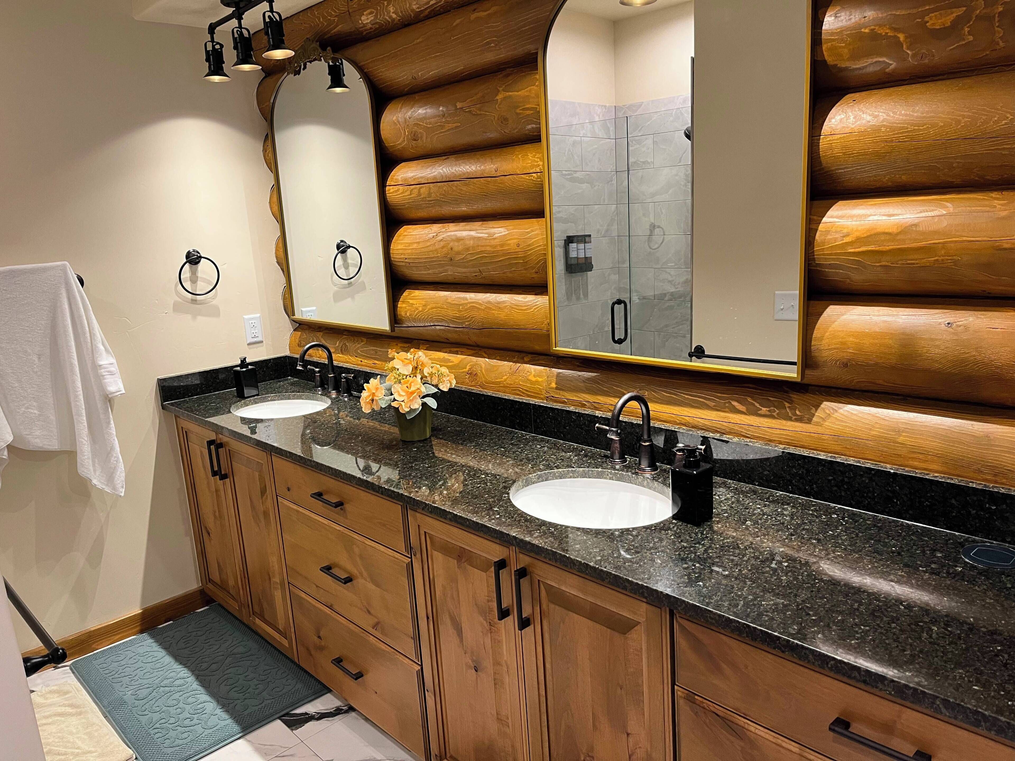 A bathroom with wooden log wall paneling, a double sink vanity, and a mirror with a towel hanging on the wall.