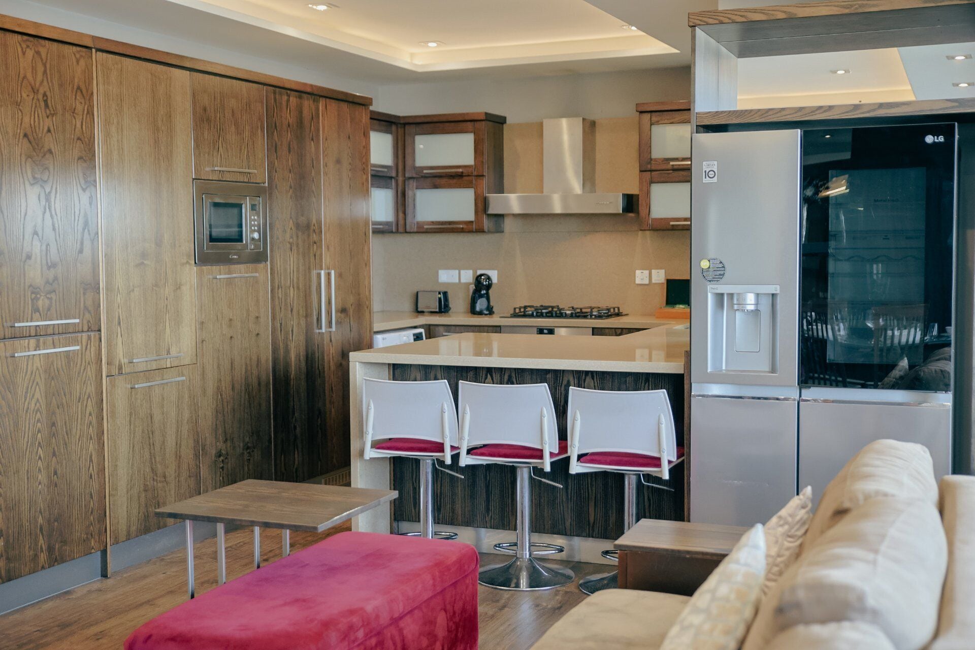 Une cuisine moderne avec des meubles en bois, des appareils en acier inoxydable, un coin bar avec des tabourets blancs et un espace détente avec un pouf rouge.