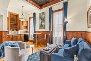 A living room with a sofa, armchair, and coffee table. There is a kitchen area with wooden cabinets and a sink.