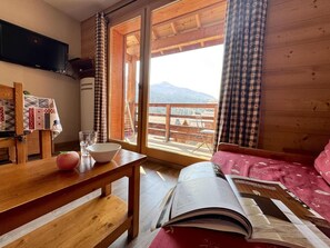 A cozy room with a wooden table, a bowl, and a book, a window with a view of mountains, and a bed with a red cover. - 50m from ski slopes, 4th floor, swimming pool, balcony, ski locker, 50m², Vars (Vars)