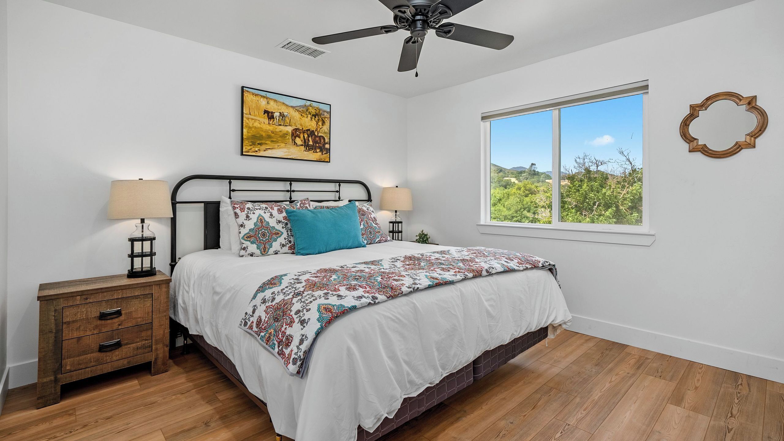 A bedroom with a bed, nightstands, a ceiling fan, a window with a view, and a painting on the wall.