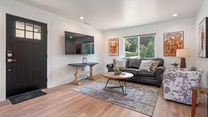 A living room with a dark door, a flat-screen TV, a coffee table, a sofa, a patterned rug, and artwork on the walls. - Stampede Loft at The 8 Mile by Avantstay Private Porch + Full Kitchen Ranch Acreage + Trails (Santa Margarita)