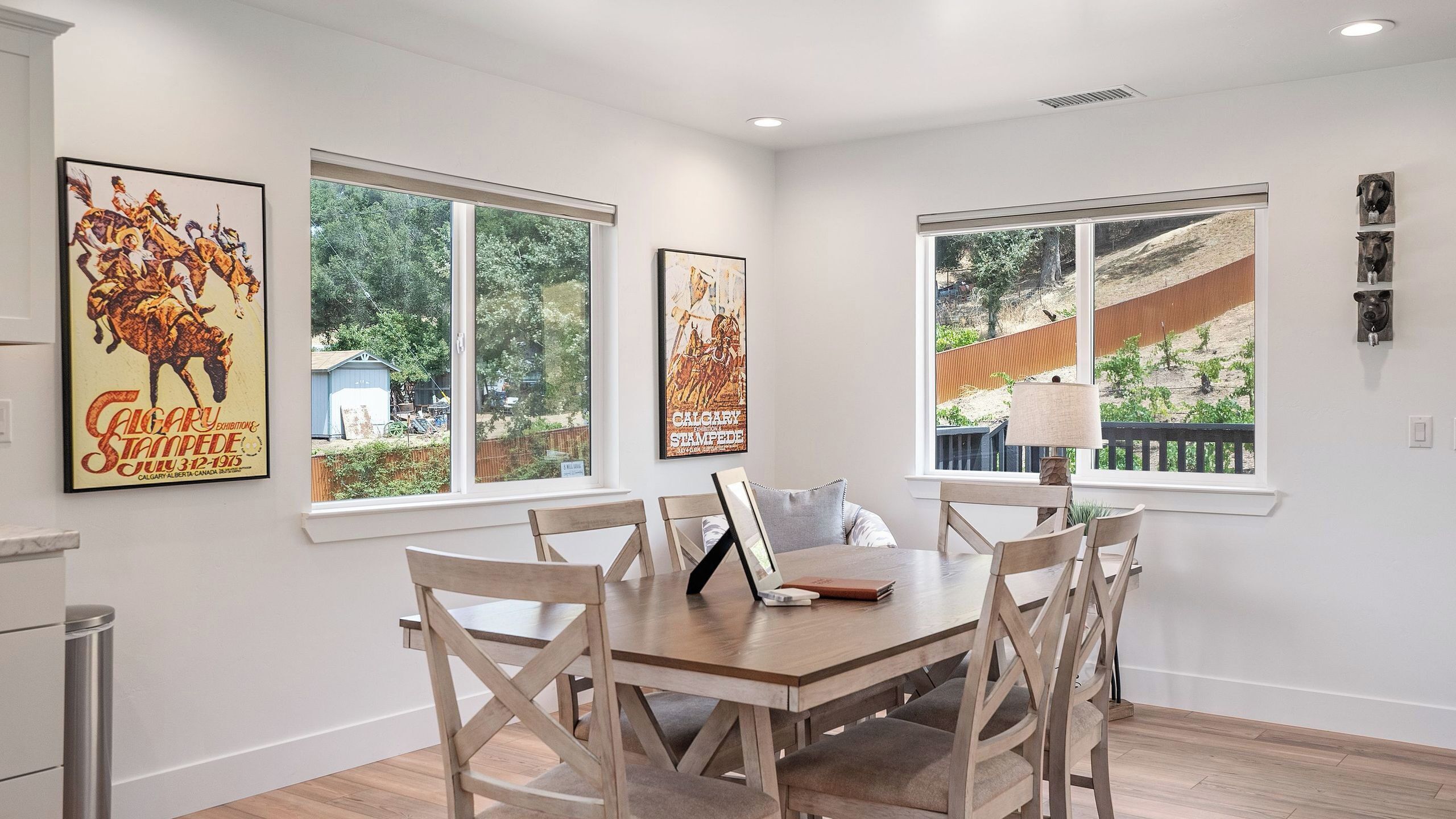 Comedor con una mesa y sillas de madera, ventanas grandes y dos cuadros colgados en la pared.