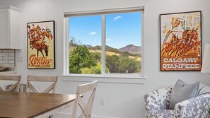 A dining area with a window view, two posters on the wall, and a patterned sofa. - Stampede Loft at The 8 Mile by Avantstay Private Porch + Full Kitchen Ranch Acreage + Trails (Santa Margarita)
