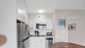 A compact kitchen with white cabinets, stainless steel appliances, and a dining area with a wooden table. - Beachfront Getaway w/ Pools, Sauna & Ocean Views (Gulf Shores)