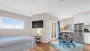 A compact room with a bed, a small table with a lamp, a kitchenette, and a TV mounted on the wall. - Beachfront Getaway w/ Pools, Sauna & Ocean Views (Gulf Shores)
