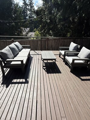 A wooden deck with outdoor furniture, including sofas and a table, surrounded by a wooden fence and trees. - Charming Indianola home, Walk to Beach, & Pier (Indianola)