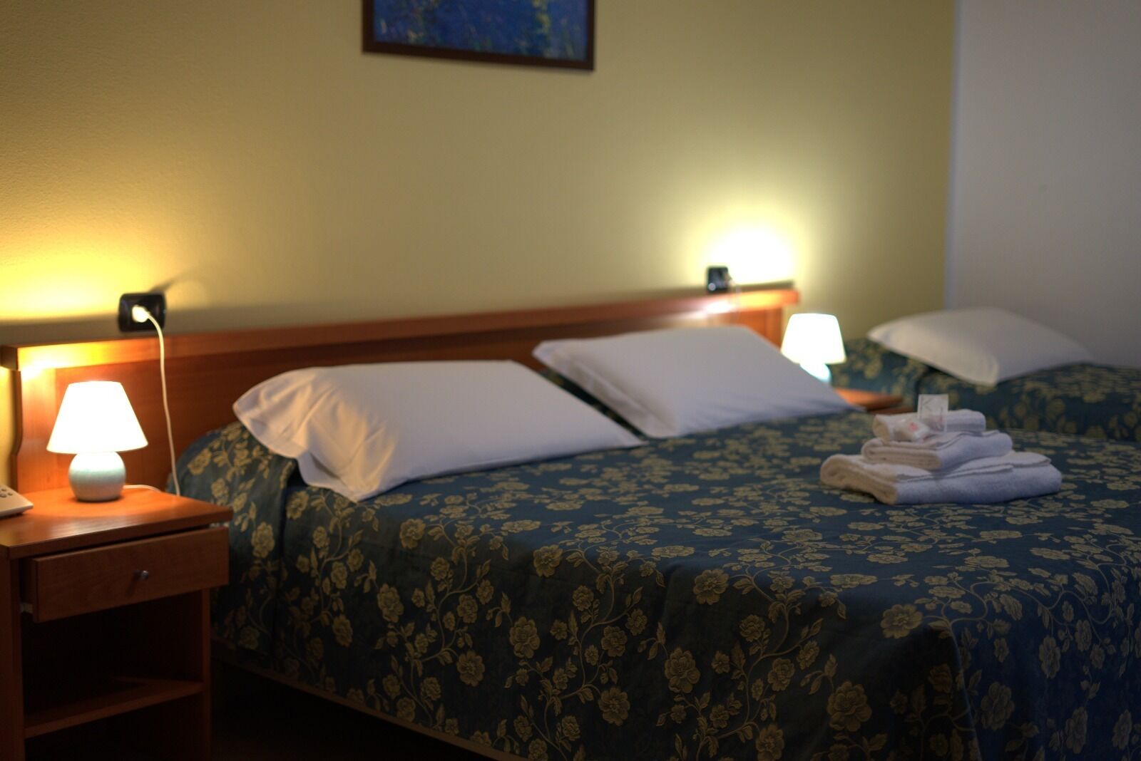 A hotel room with two beds, each with a floral bedspread and white pillows. There are bedside lamps and a folded towel on the bed.