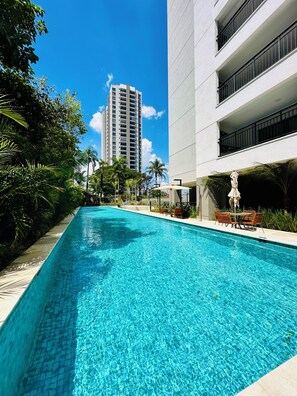 Una piscina limpia con borde de azulejos, rodeada de follaje y un edificio moderno con balcones.