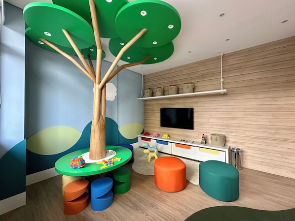 A modern children's play area with a tree-shaped play table, colorful stools, and a mounted television. - Haven Home Studio 05112 · GRU Airport + Parking + Leisure (Guarulhos)