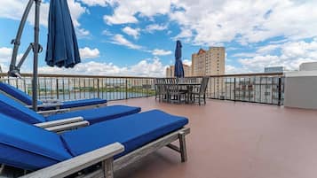 A rooftop terrace with lounge chairs and a table, overlooking a body of water and buildings.
