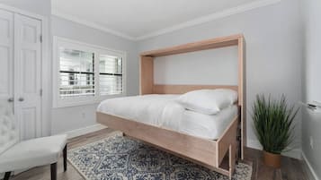 A bedroom with a bed, a chair, a patterned rug, a window, and a plant.