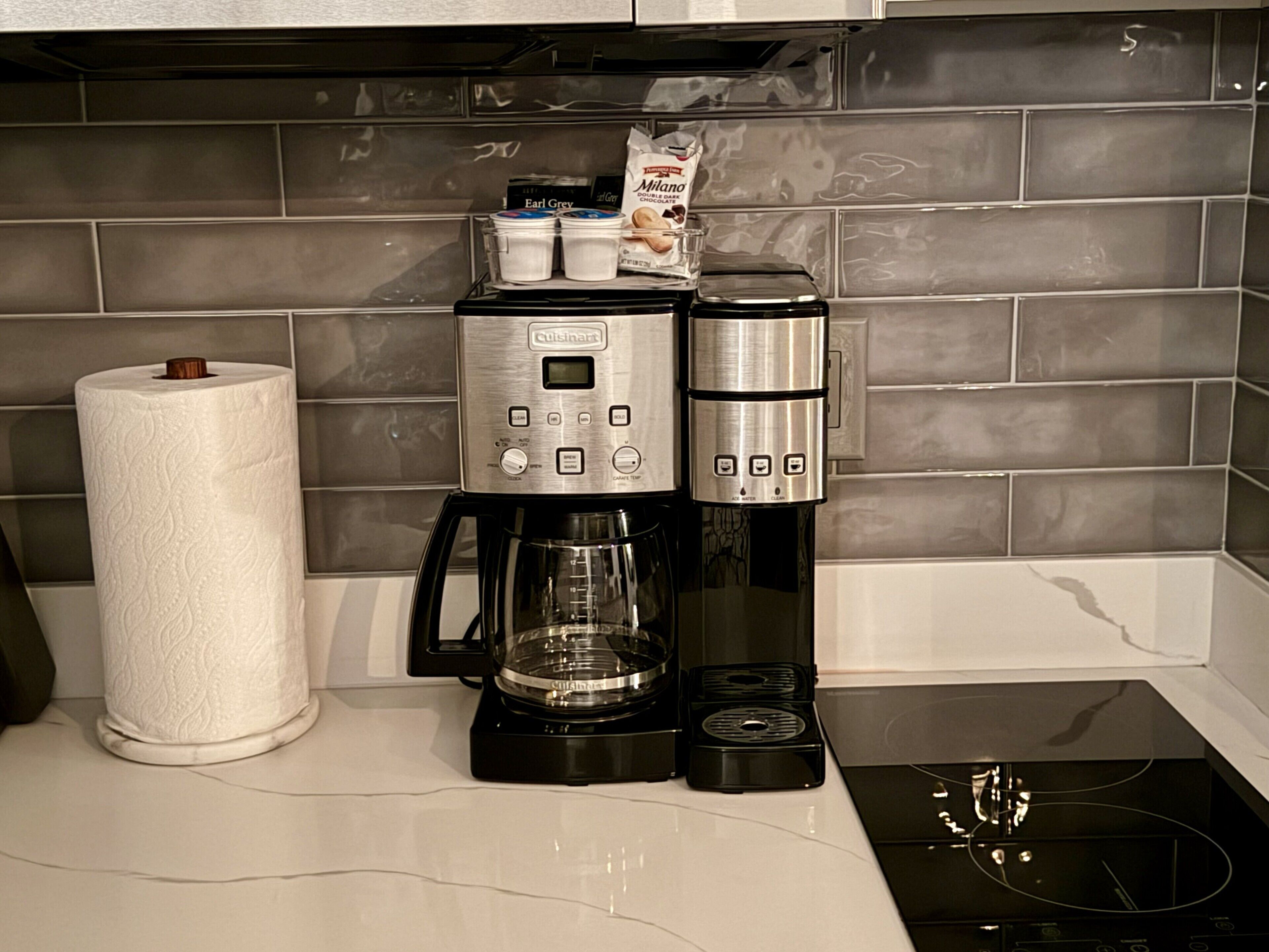 Una cafetera con pantalla digital y botones, un dispensador de toallas de papel y una jarra de café sobre una encimera de cocina.