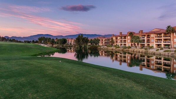 A golf course with a water body reflecting buildings, palm trees, and mountains in the background. - Coachella2ndWeek: Cozy 1-bedroom, 2 bed condo in Palm Desert:AC,resort amenities (Palm Desert)