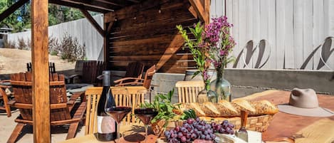 An outdoor wooden table with a bottle of wine, glasses, a vase with flowers, a basket of bread, grapes, cheese, and a knife.