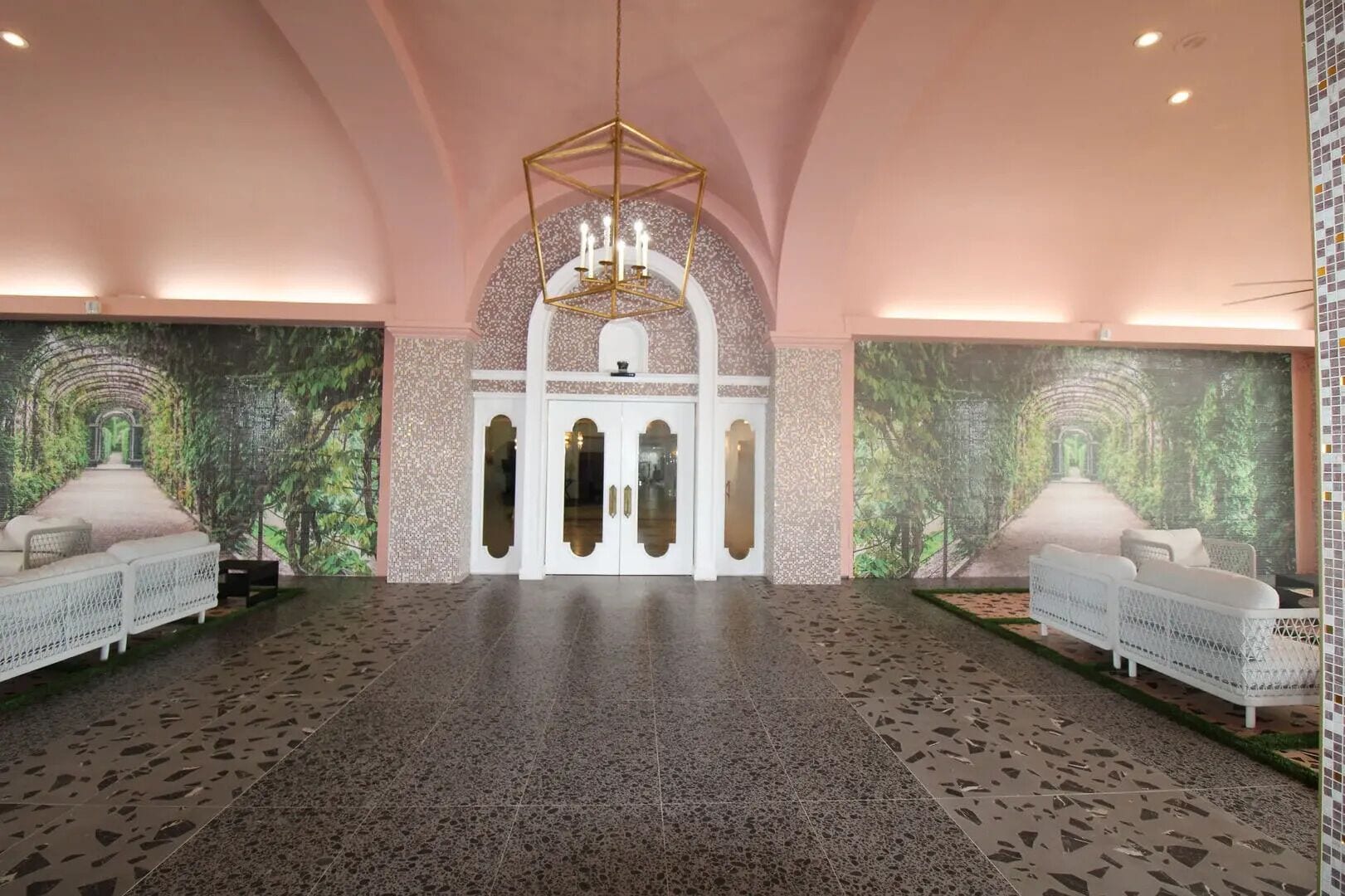 Um hall amplo com teto alto, um lustre central e duas áreas de estar com sofás brancos.