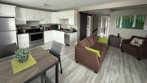 A modern kitchen with white cabinets, stainless steel appliances, and a dining area with a brown sofa and a table with a potted plant. - Executive Guest 45N (North Glengarry)