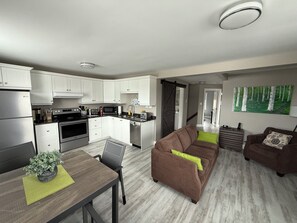 Een moderne keuken met witte kasten, roestvrijstalen apparatuur, en een zithoek met een bruine bank en een tafel met een potplant.