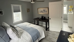 A bedroom with a bed, a desk, a ceiling fan, a window with blinds, and a door leading to another room.