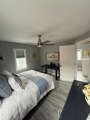A bedroom with a bed, a desk, a ceiling fan, a window with blinds, and a door leading to another room.