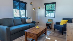 Ein modernes Wohnzimmer mit einer Couch, einem Couchtisch und einem Sessel. Es gibt zwei Fenster mit Jalousien, eine Stehlampe und einen kleinen Tisch mit einem Radio.