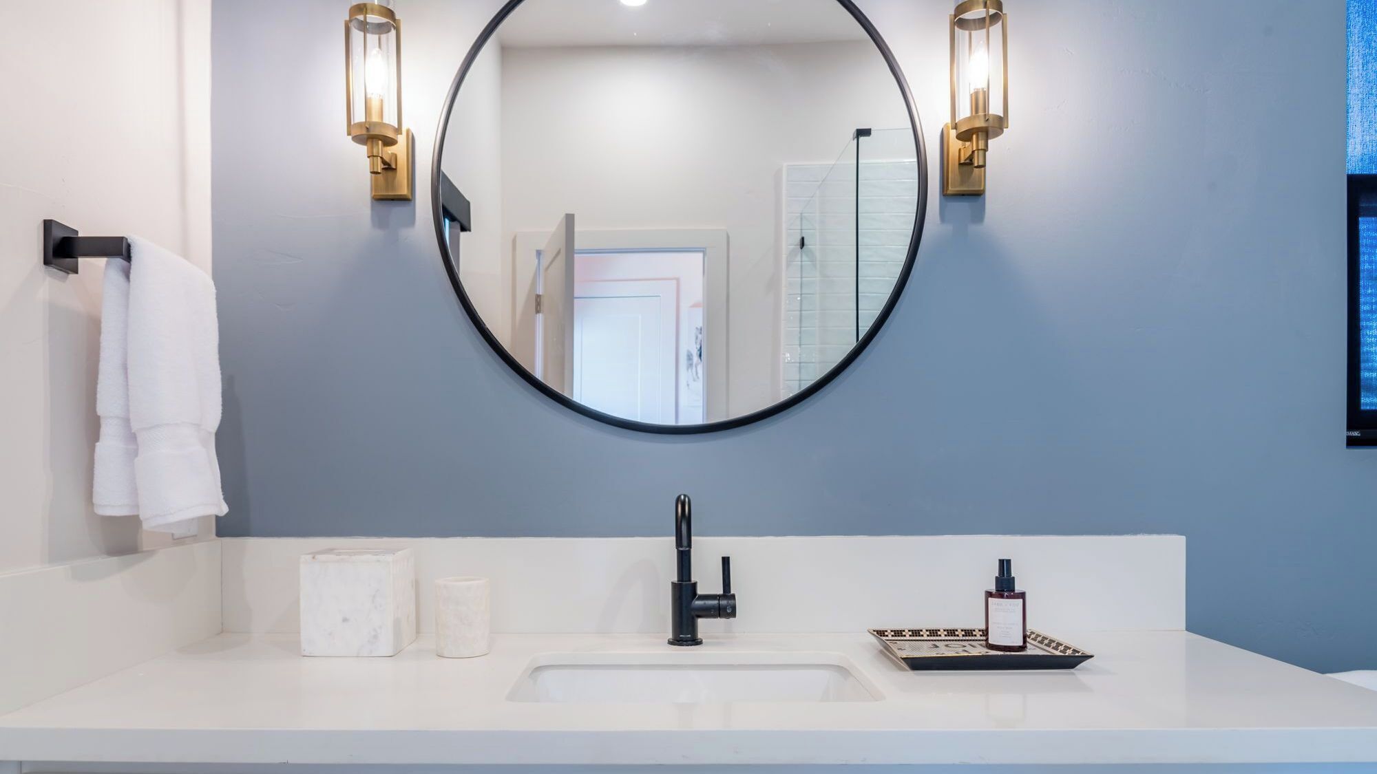 A bathroom with a round mirror, two wall-mounted lights, a white sink, a black faucet, a marble soap dish, and a bottle of shampoo.