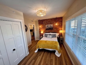 A bedroom with a wooden floor, a bed with a yellow blanket, a wall-mounted bicycle artwork, and a window with blinds. - Monroe Lofts Suite 1 Downtown 2BR Apartment (Green Bay)