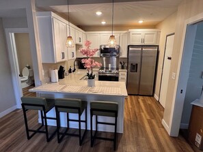 A modern kitchen with white cabinets, a stainless steel refrigerator, a central island with bar stools, pendant lighting, and a potted plant. - Monroe Lofts Suite 1 Downtown 2BR Apartment (Green Bay)