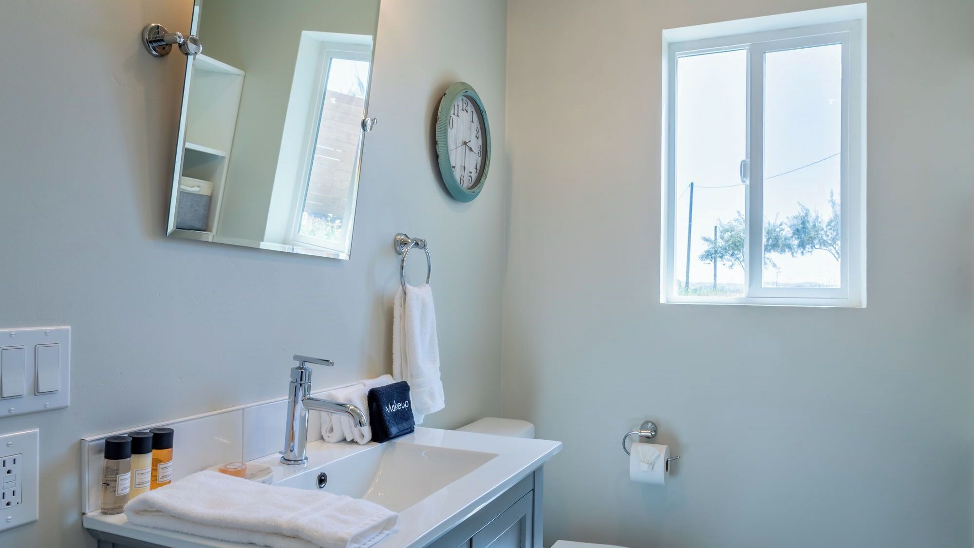 Une salle de bain avec un lavabo blanc, un miroir, une horloge, un porte-serviettes et une fenêtre donnant sur des arbres.