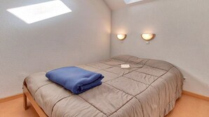 Une chambre avec un puits de lumière au plafond, deux lampes fixées au mur, un lit avec une couette beige et un oreiller bleu, et une petite table de chevet.