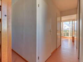 Un pasillo con una puerta de madera a la izquierda, una puerta blanca en el centro y vista a un balcón con una montaña al fondo.