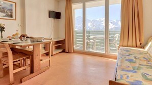 Une pièce comprenant un lit, une table avec des chaises, un téléviseur fixé au mur et une grande fenêtre avec des rideaux.