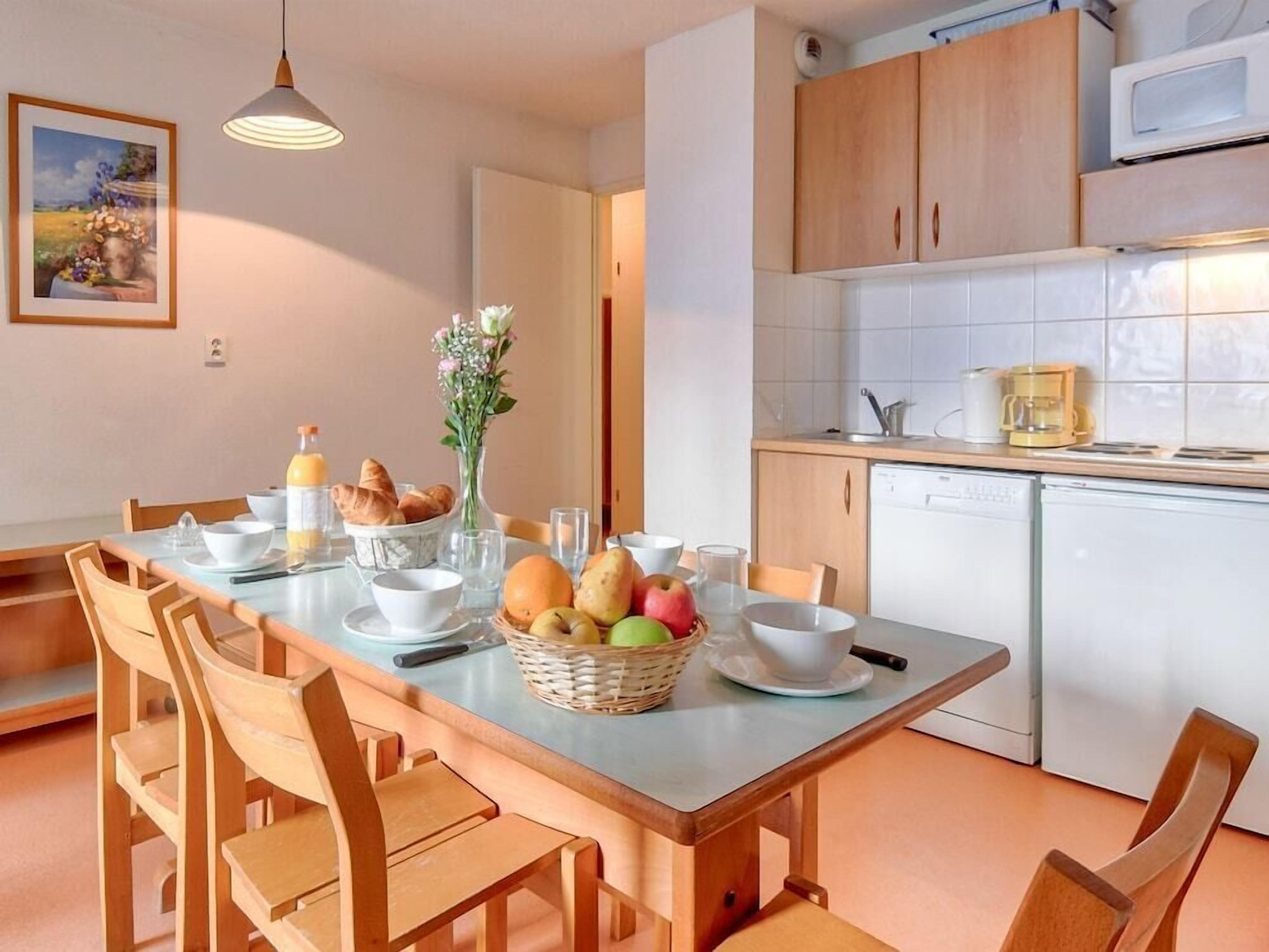 Un coin repas avec une table dressée pour deux personnes, une cuisine dotée d’appareils électroménagers blancs et d’armoires en bois.