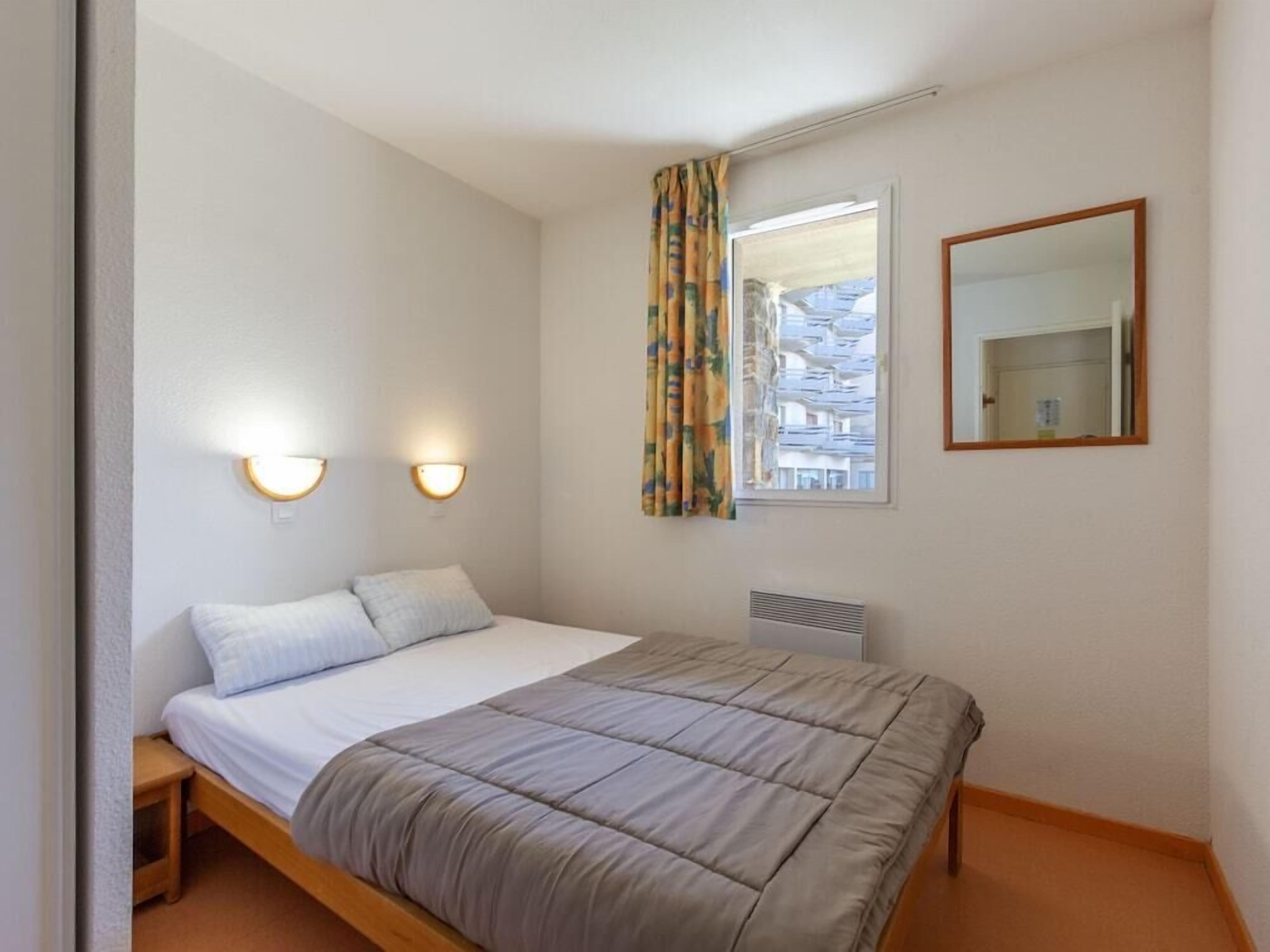 Une chambre à coucher avec un lit, deux lampes fixées au mur, une fenêtre avec des rideaux et un miroir.