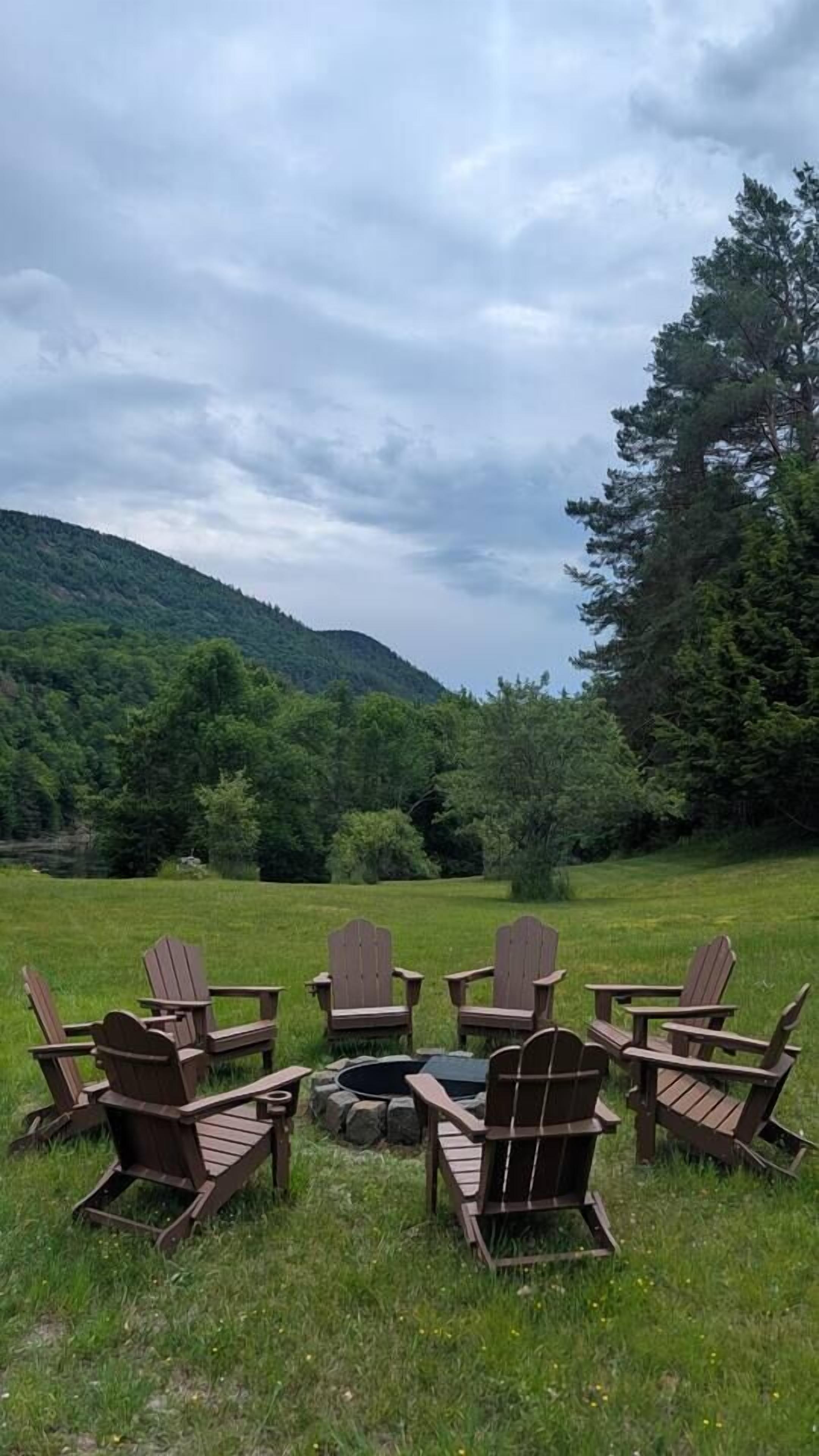 A group of Adirondack chairs arranged around a fire pit in a grassy field with a forested hill in the background.