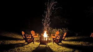 A campfire with two Adirondack chairs facing it, set on a grassy area at night. - Cabin with Private Pond - 61 Acres - Close to Gore (Johnsburg)