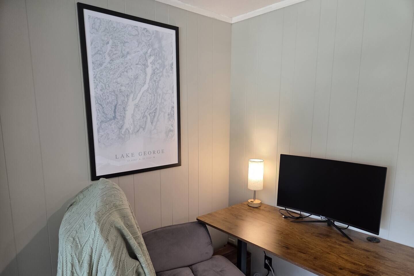 A room with a framed map on the wall, a flat-screen TV on a wooden table, a lamp, and a grey sofa.