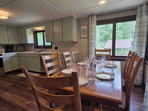 A dining area with a wooden table set for six, surrounded by wooden chairs. A kitchen is visible in the background with light green cabinets. - Cabin with Private Pond - 61 Acres - Close to Gore (Johnsburg)