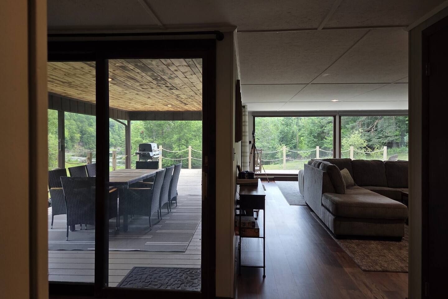 Een modern interieur met een houten terras, buitenmeubelen en een bank tegenover grote ramen.