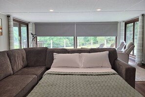 A room with a large sectional sofa, a bed with a patterned bedspread, and a view of the outdoors through large windows. - Cabin with Private Pond - 61 Acres - Close to Gore (Johnsburg)
