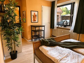 Ein Zimmer mit einem Bett, einem kleinen Tisch, einer Topfpflanze und einem Fenster mit Blick ins Freie.