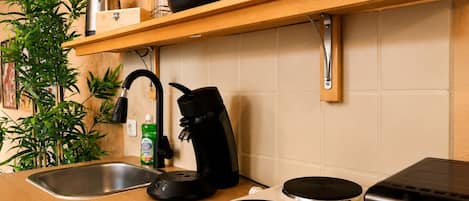 A kitchen with a stainless steel sink, a kettle, a coffee maker, and a microwave. There are wooden shelves above the counter holding plates and a bottle.